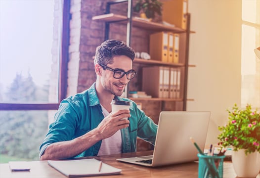 Hombre joven con un vaso de café en la mano, trabajando frente a su computador portátil.