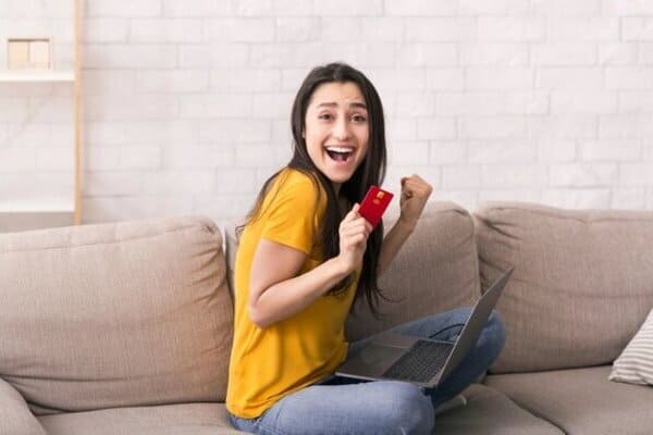 Mujer joven feliz, sentada en la sala de su casa, realizando compras con su tarjeta de crédito.