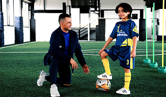 Niño y entrenador practicando fútbol en una cancha sintética.