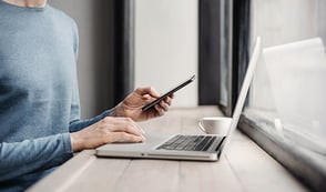 Persona con su teléfono en la mano, trabajando en su computador portátil.