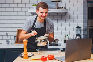 Hombre con una cuchara y una olla atiende exposición virtual desde su computador portátil.