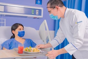 Enfermero lleva comida a mujer que reposa en cama hospitalaria.