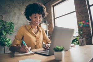 Mujer joven frente a su computador portátil, tomando apuntes en su agenda.