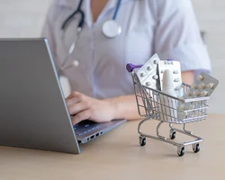 Una médica trabajando su computador portátil y a lado un carrito de compras a escala pequeña con pastillas.