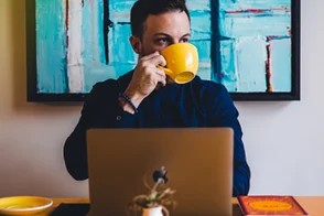 Hombre joven, frente a su computador portátil, tomando una taza de café.