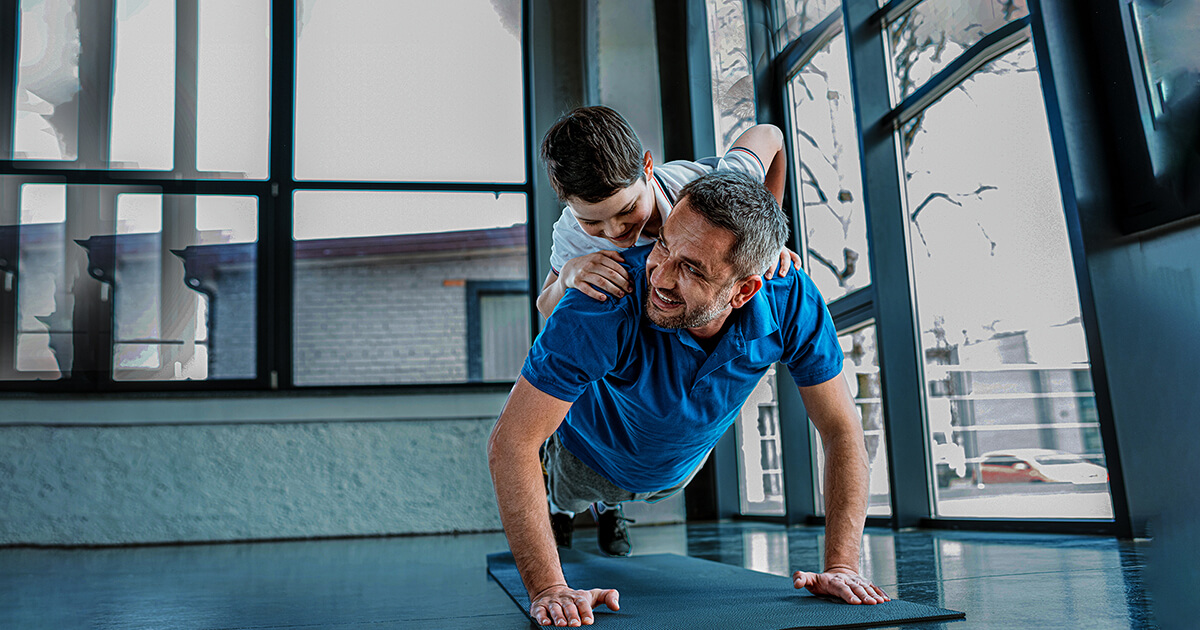 Padre entrenando en el gimnasio Colsubsidio y su hijo encima de su espalda. 