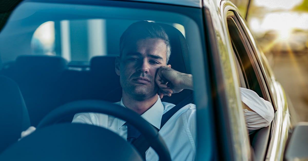 Hombre conduciendo su vehículo, quien tiene expresión de aburrimiento y molestia por los trancones. 