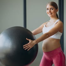 Una mujer embarazada levanta con sus manos un balón de yoga mientras sonríe.